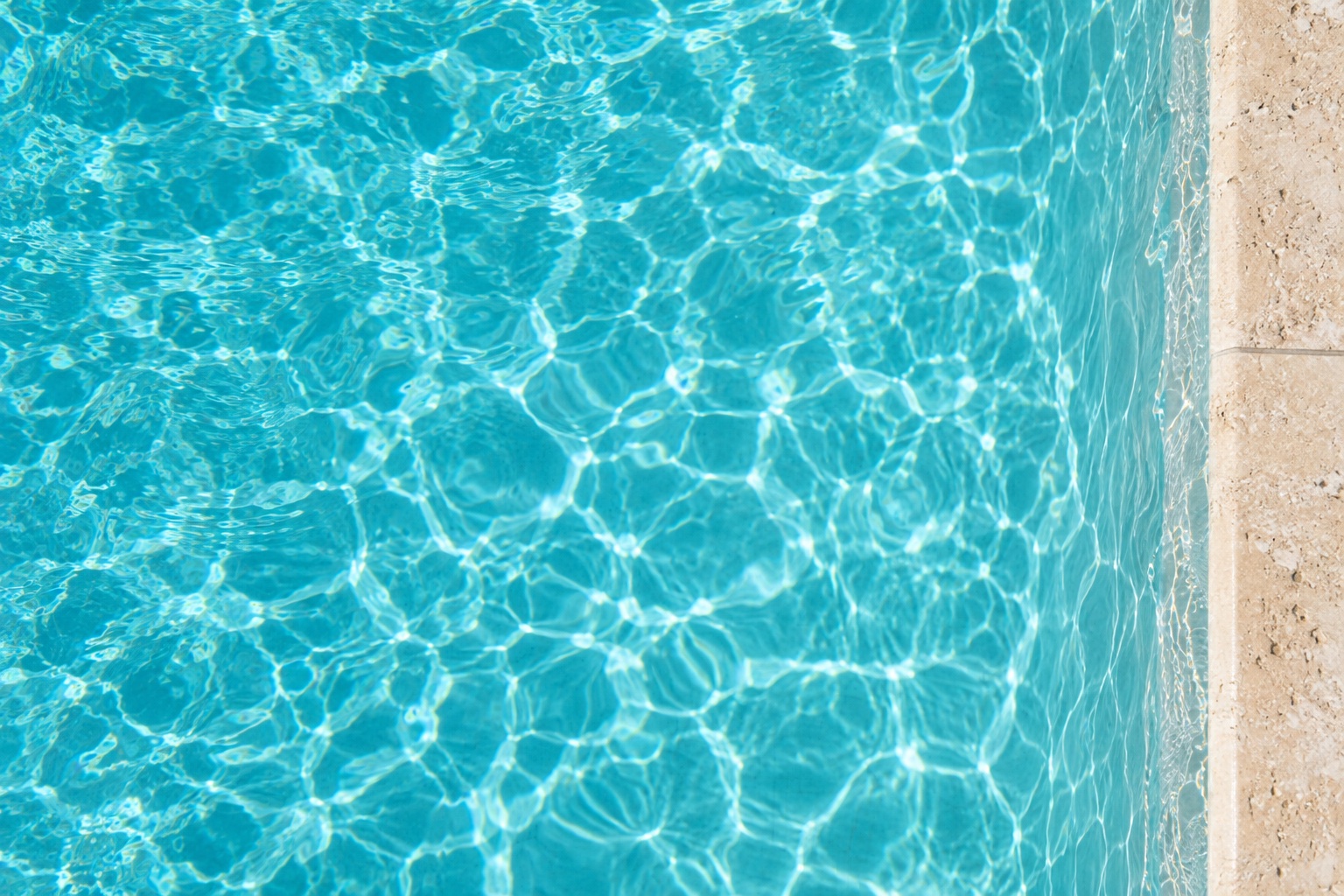 Sunlight moving across turquoise pool water beside pale stone coping